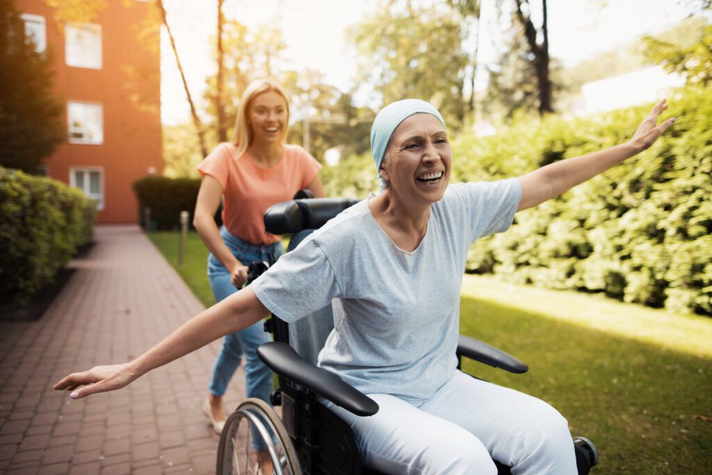 A woman with cancer is sitting in a wheelchair. She walks on the street with her daughter and they fool around. They are fun and they laugh. They walk in the courtyard of the clinic.